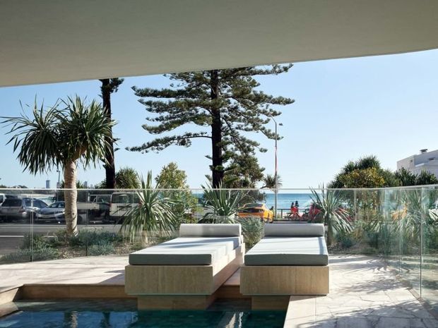 A bright, contemporary outdoor lounge beside a swimming pool, with two low upholstered daybeds on timber-clad plinths. Glass balustrading separates the pool terrace from a landscaped coastal streetscape and ocean beyond. The space is sheltered by a broad overhead roof or soffit and surrounded by palms and sculptural planting.
