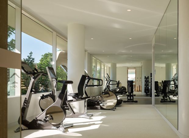 A bright, elongated home gym with modern cardio and strength-training equipment arranged along floor-to-ceiling windows. Full-height mirrored panels line the right wall, reflecting the machines and creating a sense of spaciousness. The room has white cylindrical columns, recessed ceiling lights, pale carpet, and a clean, refined contemporary fit-out with abundant natural light.