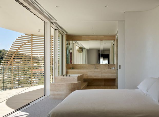 Bright contemporary bedroom with a low-profile bed, full-height sliding glass doors opening to a balcony, and a sculptural timber privacy screen outside. The room features warm natural timber or stone-look built-in vanity and cabinetry, a large mirrored wall, pale carpet, white walls, and a clean minimalist fit-out.