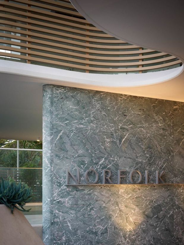 A sophisticated hotel or commercial lobby featuring a dramatic curved reception feature wall clad in veined grey-green natural stone, with raised metallic lettering reading “NORFOLK.” Above, a sweeping white architectural soffit and layered timber-look slatted ceiling create a sculptural canopy. Floor-to-ceiling glazing and indoor planting are visible at the side, with warm integrated lighting washing the stone wall.