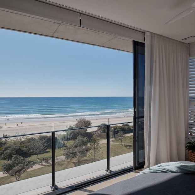 Bright coastal bedroom interior with a bed in the foreground, full-height sliding glass doors, sheer curtains, and a balcony overlooking a sandy beach, ocean, landscaped foreshore, and walking path. The space has a clean contemporary feel with strong indoor–outdoor connection and expansive ocean views.