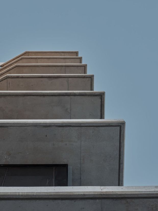 Upward-looking architectural photograph of a stark stepped concrete facade composed of projecting rectangular volumes and parapets, with a small dark window opening at the lower left. The raw grey concrete is set against a clear muted blue sky, creating a minimalist geometric composition.