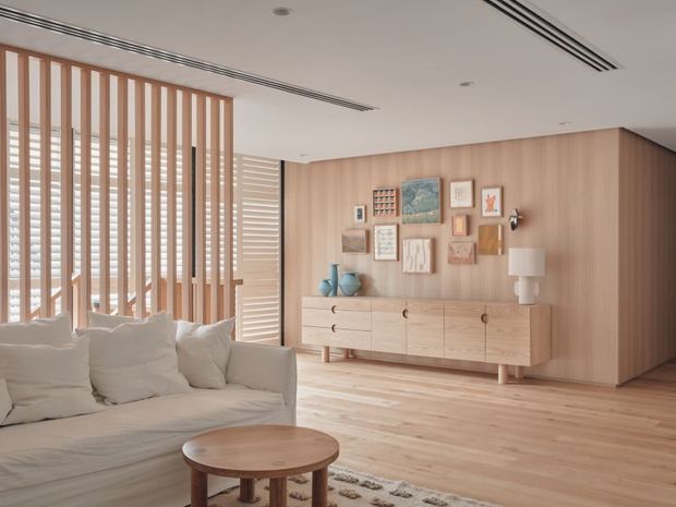 Bright contemporary living room with a pale timber-lined feature wall and matching built-in sideboard, a light upholstered sofa, round timber coffee table, vertical timber screening, plantation shutters, and a curated arrangement of framed artwork. The room has a clean, warm and airy appearance with recessed linear air vents and subtle decorative lighting.