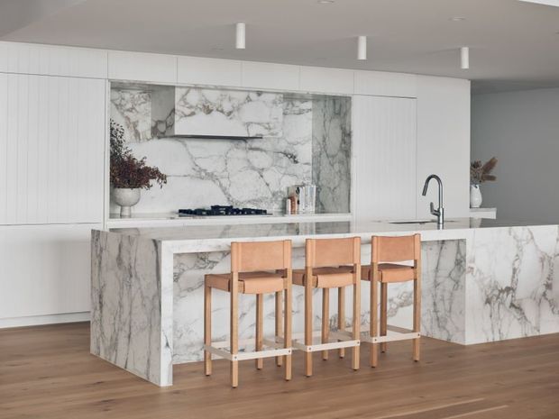 A bright, contemporary kitchen featuring extensive white cabinetry, a dramatic white-and-grey veined stone splashback and island, integrated cooktop, and a minimalist dark metal tap. Four timber bar stools line the island, contrasting with the warm timber flooring. The space has a clean, refined and highly cohesive material palette.