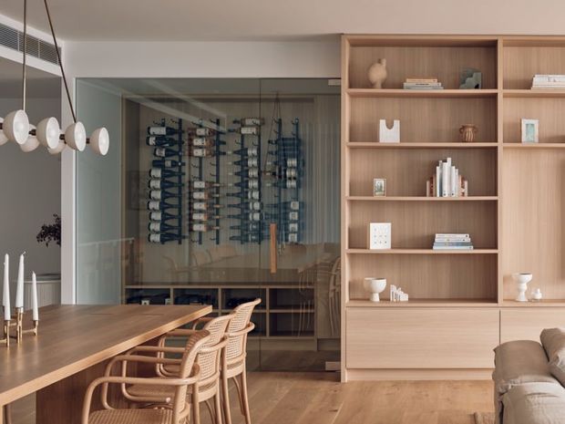 Warm contemporary dining and living area with a long timber dining table, woven rattan dining chairs, sculptural pendant lights, and a large built-in timber shelving unit. A glass-fronted wine display and storage wall forms a feature behind the dining area, while part of a neutral sofa is visible in the foreground.