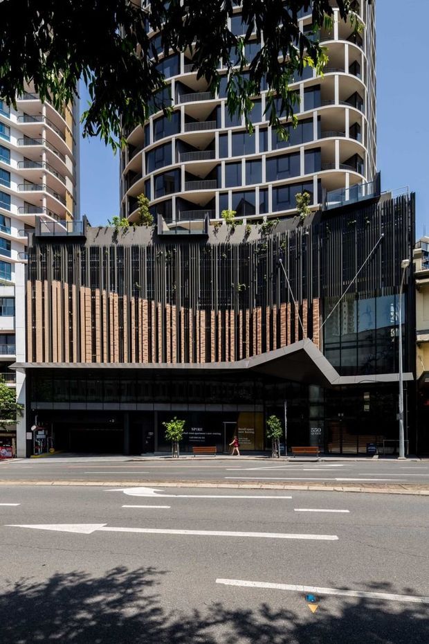 Street-level view of a contemporary multi-storey urban apartment and commercial building. The facade combines curved residential balconies, extensive dark glazing, vertical black metal screening, warm timber-toned slats, and a projecting angular canopy over the ground-floor entry. A broad city street and neighbouring high-rise buildings are visible.