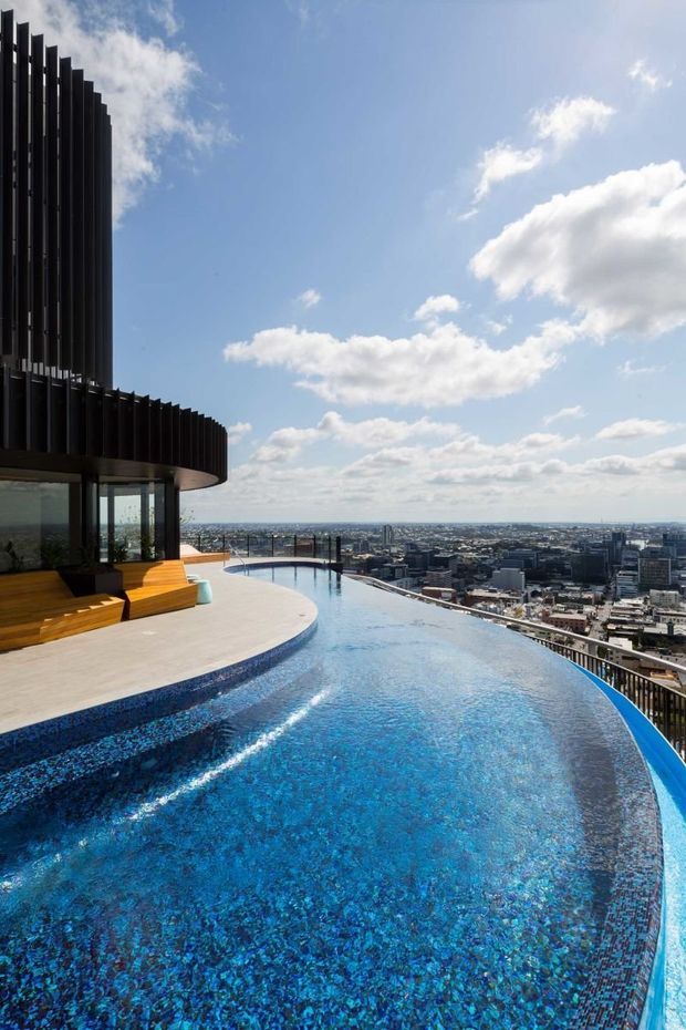 A rooftop infinity pool overlooks a dense urban skyline beneath a bright blue sky. The pool has vivid blue mosaic tile lining and a curved edge, with a pale paved deck, built-in timber seating, glass balustrades, and a dark vertical louvred architectural facade wrapping the upper left side.
