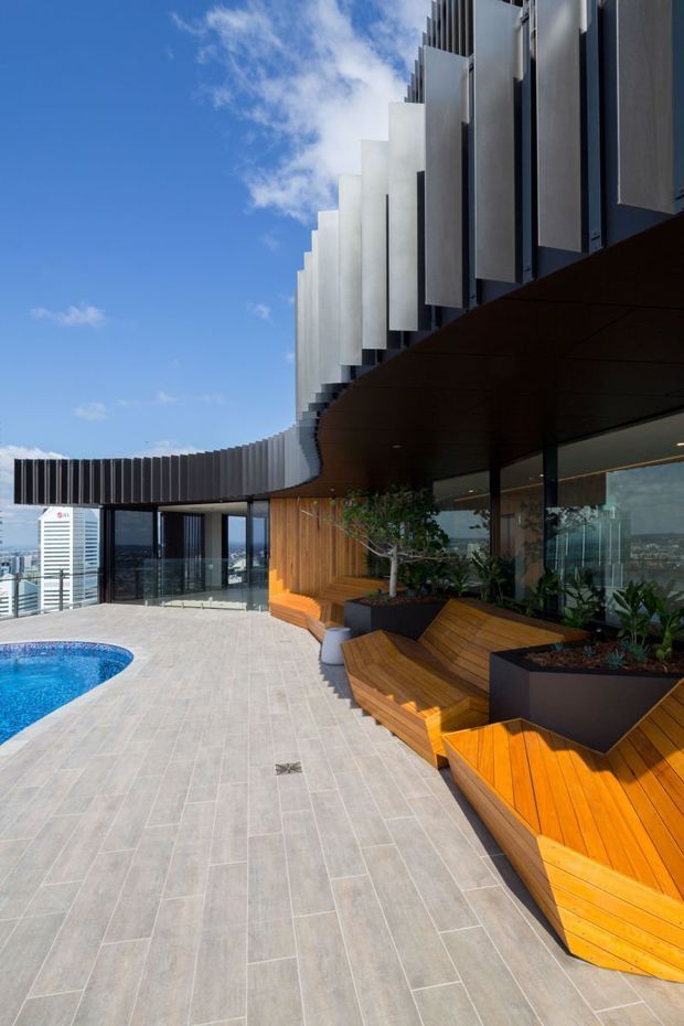 A rooftop terrace with a curved swimming pool, pale tiled paving, built-in timber seating and planters, and a contemporary building facade with projecting vertical fins and extensive glazing. The terrace overlooks a dense urban cityscape under a bright blue sky.