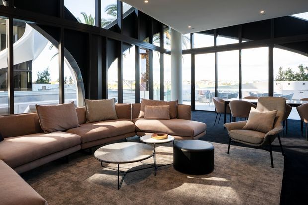 A spacious contemporary lounge and dining area with floor-to-ceiling black-framed glazing, abundant natural light, and views of palm trees and neighboring buildings. The seating area features a large modular taupe sectional, upholstered armchairs, nested round coffee tables, a dark cylindrical side table, and a broad neutral rug. A dining table and chairs sit near the windows, while a striking curved white architectural element is visible outside.