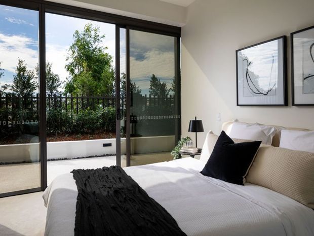 Bright contemporary bedroom with a neatly made bed, white bedding, a black accent cushion and throw, bedside table and lamp, and abstract framed artwork. Full-height dark-framed sliding glass doors open to a planted outdoor terrace or balcony, bringing in abundant natural light.