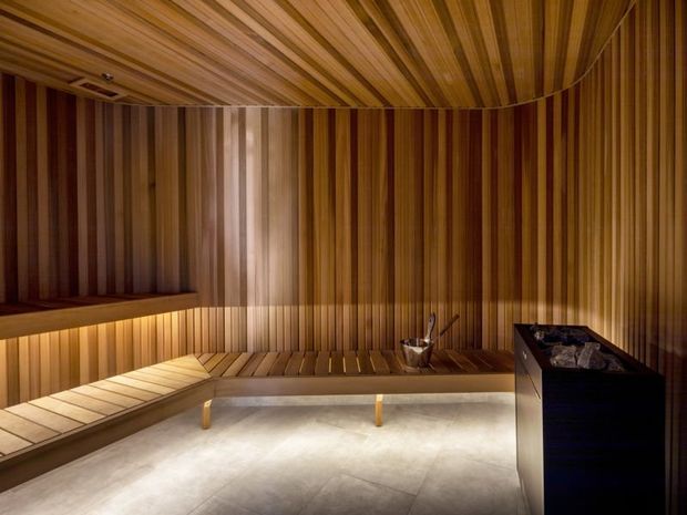 A compact, warmly lit sauna clad throughout in vertical and linear timber slats. Built-in timber benches wrap around the room, with a dark sauna heater at the right and a pale smooth floor below. The strongly continuous timber ceiling and walls create a refined, minimalist spa-like interior.