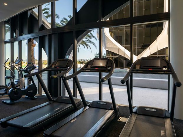 A modern gym interior featuring three treadmills in the foreground and additional cardio equipment to the left. Floor-to-ceiling black-framed glazing opens views toward palm trees and a striking curved white architectural structure outside. The space has a dark, polished, contemporary fit-out with strong natural light and high visual contrast.