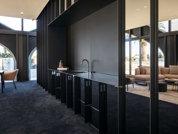 A sophisticated open-plan interior featuring a long black built-in kitchenette or bar cabinetry with a dark benchtop, integrated sink and curved tap. Dramatic black vertical wall panelling and structural framing divide the space, while floor-to-ceiling glazing opens views to a bright outdoor setting. A lounge area with neutral upholstered seating is visible beyond the partition, and dark carpeting covers the floor.