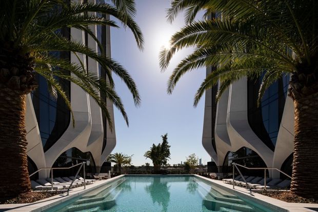 Symmetrical exterior view of a landscaped resort-style pool courtyard between two sculptural contemporary buildings. Tall palm trees frame the turquoise rectangular swimming pool, with curved white and dark glazed facades, sun loungers, and stainless-steel pool rails visible on both sides.