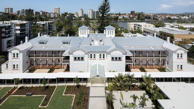 Aerial view of a large, symmetrical heritage-style institutional or residential building with pale rendered walls, an extensive light-grey metal roof, multiple small cupola-like towers, arched verandahs, and landscaped formal gardens. Contemporary apartment and commercial buildings, a waterway, and an urban skyline are visible in the background.