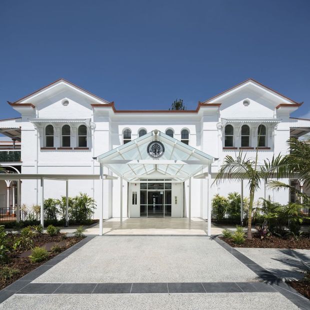 Front elevation of a symmetrical white two-storey building with gabled wings, classical arched windows, decorative white detailing, and a glazed entrance canopy. A paved forecourt and tropical landscaping lead to the central entry.