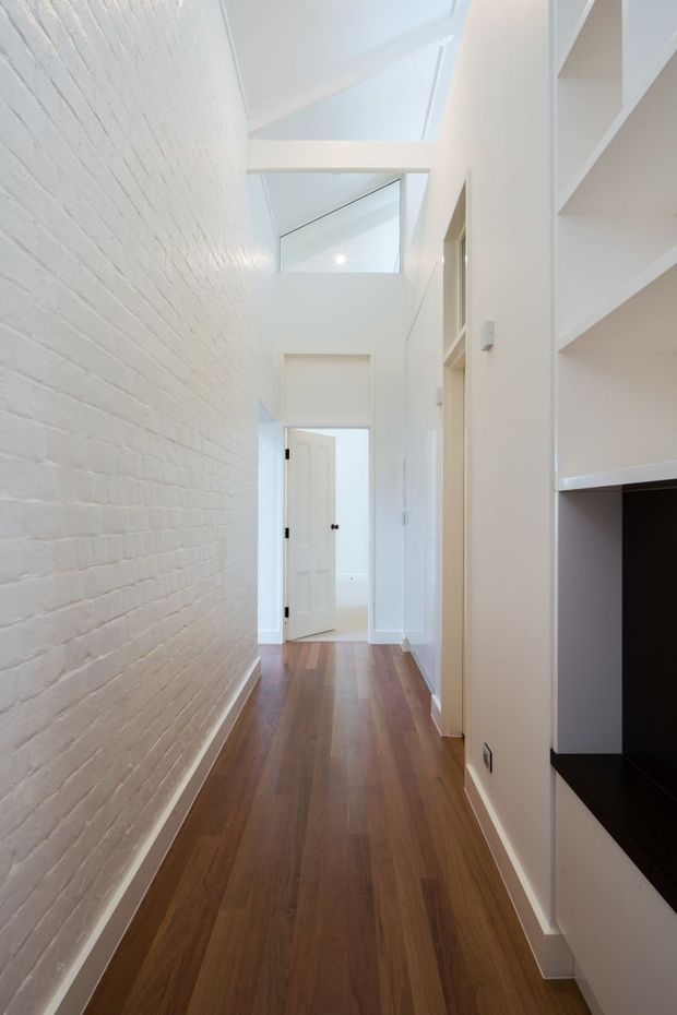 Bright, narrow residential hallway with a warm timber floor, white-painted exposed brick wall, white plastered walls and built-in open shelving. A glazed high-level opening and skylight-like roof glazing bring abundant natural light into the corridor, with a white door visible at the far end.