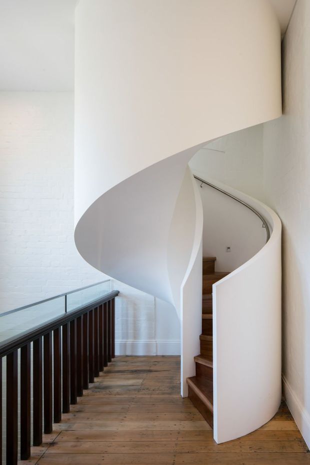 A sculptural curved staircase with a smooth white spiral balustrade and enclosing form, dark timber vertical railings with a glass guardrail, and warm timber floorboards. The staircase sits against white-painted brick and plaster walls, creating a minimalist, light-filled architectural interior.