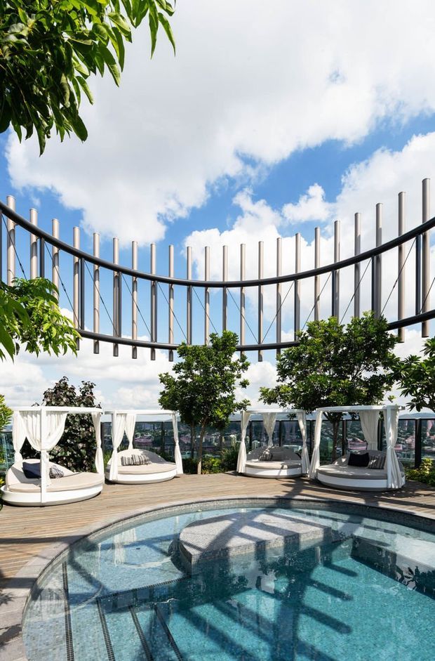 A luxurious rooftop pool terrace with curved blue-tiled swimming pool, timber decking, white curtained cabana daybeds, lush planted trees, and a dramatic suspended circular architectural structure overhead. The terrace overlooks a dense urban skyline beneath a bright partly cloudy sky.