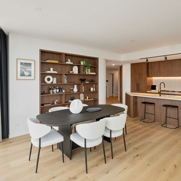 An open-plan contemporary kitchen and dining area with warm timber cabinetry, a large built-in display shelving wall, pale timber flooring, and a dark dining table surrounded by white upholstered chairs. The kitchen includes a stone benchtop, integrated warm under-cabinet lighting, black tapware, and bar stools. Neutral walls and restrained decor create a refined, welcoming interior.