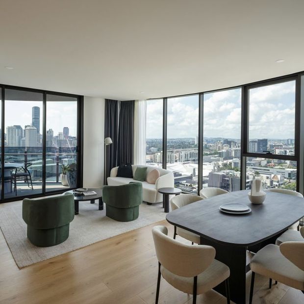 A bright, high-rise apartment living and dining area with expansive floor-to-ceiling corner glazing overlooking a dense city skyline. The room features pale timber flooring, a neutral rug, a dark oval dining table with upholstered dining chairs, rounded green lounge chairs, a cream sofa, dark curtains, and understated contemporary furnishings.