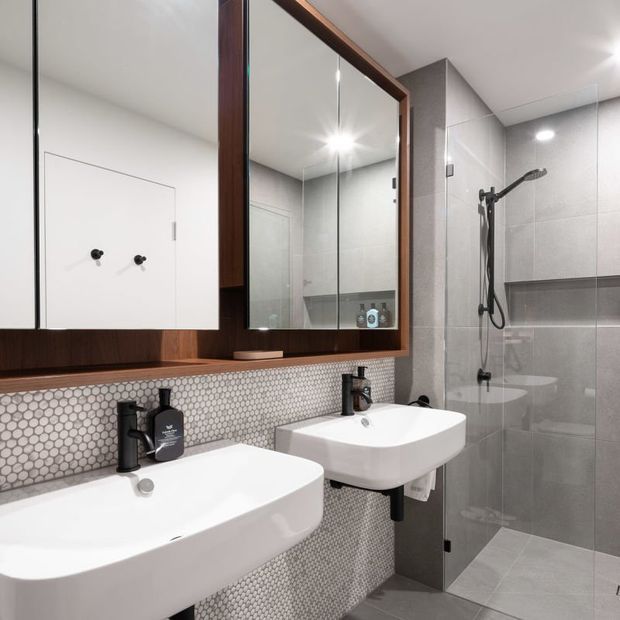 Contemporary bathroom featuring two white wall-mounted basins with matte black tapware, a large timber-framed mirrored cabinet, grey hexagonal mosaic wall tiles, and a grey tiled walk-in shower with black fittings. The compact space has clean lines, integrated storage, and bright recessed lighting.