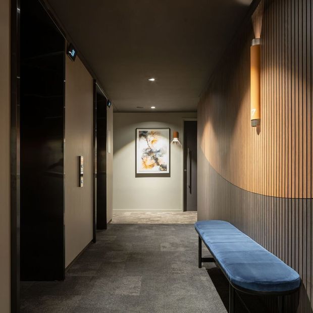 A moody, refined interior hallway with dark carpet flooring, muted painted walls, concealed doors or cabinetry along the left side, and a dramatic vertical timber slat feature wall on the right. Warm integrated wall sconces illuminate the curved feature detail, while a long blue upholstered bench provides seating. Artwork and a doorway are visible at the far end.