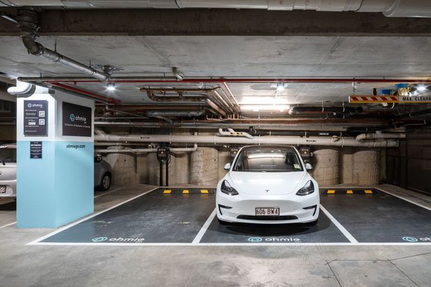 An underground commercial parking garage with marked electric-vehicle charging bays. A white Tesla is parked in a designated charging space beside an EV charging station, with exposed concrete structure, overhead services, pipes, ducts, and industrial fluorescent lighting.