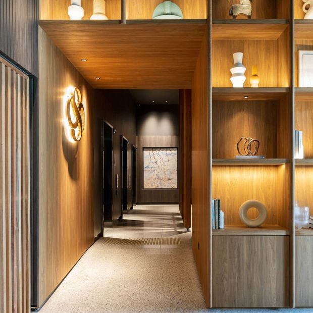 A refined interior hallway framed by warm timber wall panelling, a timber-lined ceiling, and built-in open shelving filled with decorative objects and books. The corridor features dark doorways, recessed lighting, a terrazzo-like light floor, and a sculptural wall light, creating a luxurious, carefully detailed passage.