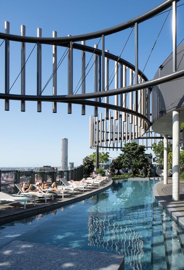 A rooftop pool terrace in a contemporary high-rise setting, featuring a long reflective swimming pool, curved dark metal architectural structures with vertical rods and tension cables, pale tiled decking, lounge chairs, landscaped planters, and panoramic urban views.