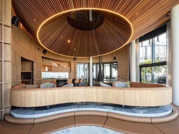A refined contemporary reception or lounge interior with extensive curved timber slat wall and ceiling panelling. A large elliptical illuminated feature pendant dominates the ceiling above a sculptural curved timber reception desk or built-in seating element, with integrated soft brown upholstered seating and a dark blue-grey tiled platform. Floor-to-ceiling glazing brings in daylight and overlooks an urban exterior. The space has a warm, luxurious, highly crafted hospitality-like atmosphere.