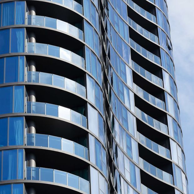 Close-up exterior view of a tall contemporary apartment building with strongly curved stacked balconies, blue-tinted floor-to-ceiling glazing, transparent glass balustrades, dark balcony soffits, and reflective metal-framed curtain-wall panels. The facade has a fluid, sculptural form against a pale sky.