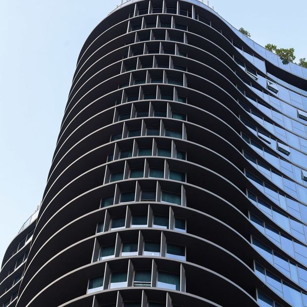 Upward-looking view of a tall contemporary multi-storey building with a rounded facade, repeated projecting balcony bands, dark vertical screens, and extensive reflective glazing. A small amount of rooftop planting is visible against a clear sky.