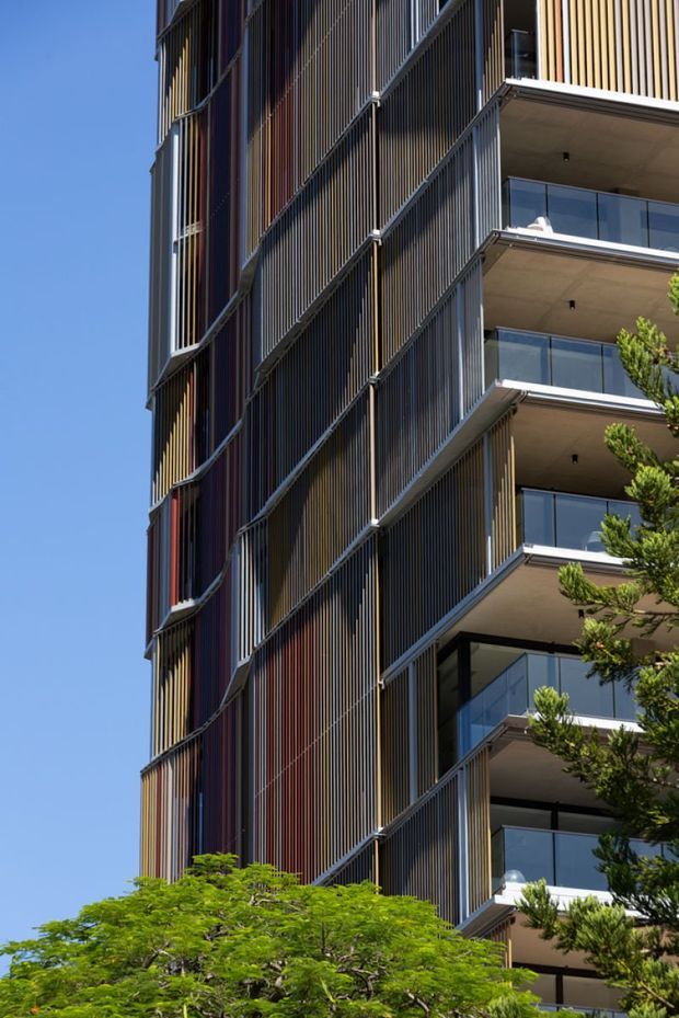Vertical view of a contemporary multi-storey apartment building with projecting balconies, transparent glass balustrades, and a prominent facade of closely spaced vertical fins in varied dark, bronze, and reddish tones. The building is framed by mature trees against a clear blue sky.