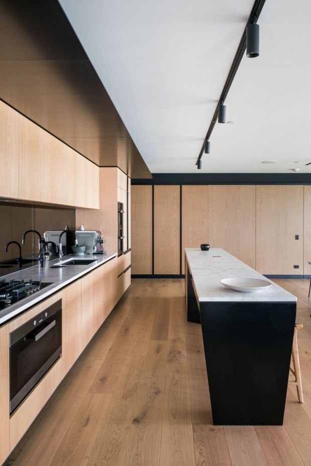 Contemporary kitchen with a long run of light timber cabinetry, integrated black appliances, a dark stone or composite benchtop, and a large contrasting island with a pale stone surface and black base. The room features timber flooring, full-height timber wall panels, black trim, and a linear track of black cylindrical spotlights.