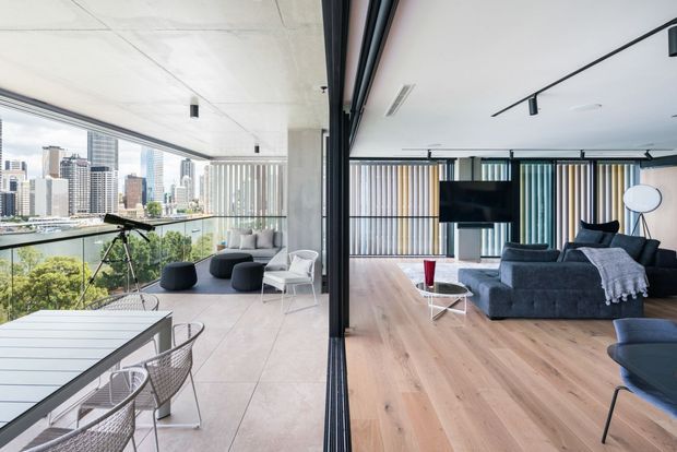 A contemporary open-plan apartment interior with a living room and dining area connected by fully retractable black-framed glazing. The room features floor-to-ceiling windows overlooking a dense city skyline and greenery, pale timber flooring, large-format light stone or concrete-look balcony flooring, exposed concrete ceiling and column elements, a charcoal sectional sofa, minimalist furniture, and vertical striped window blinds. A dining table and wire-frame chairs occupy the glazed terrace or balcony area.