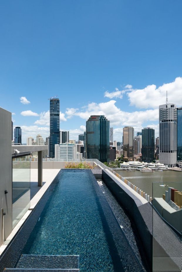 A rooftop infinity-style swimming pool with dark blue mosaic tiling, glass balustrades, and a clean modern terrace overlooking a dense high-rise city skyline and waterfront marina under a bright blue sky.