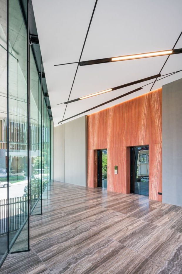 A tall, contemporary interior lobby or entry space with full-height glazed walls, a dramatic veined stone floor, a warm reddish stone feature wall surrounding dark-framed doorways, and suspended linear lighting arranged across the ceiling. The space is largely open and minimally furnished, with views to an exterior landscaped area through the glazing.