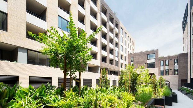 Contemporary multi-storey apartment or mixed-use development arranged around a landscaped communal courtyard. The facade uses pale brick, recessed balconies, dark-framed glazing, and strong repetitive vertical window bays, with lush garden planting and paved pedestrian circulation at ground level.
