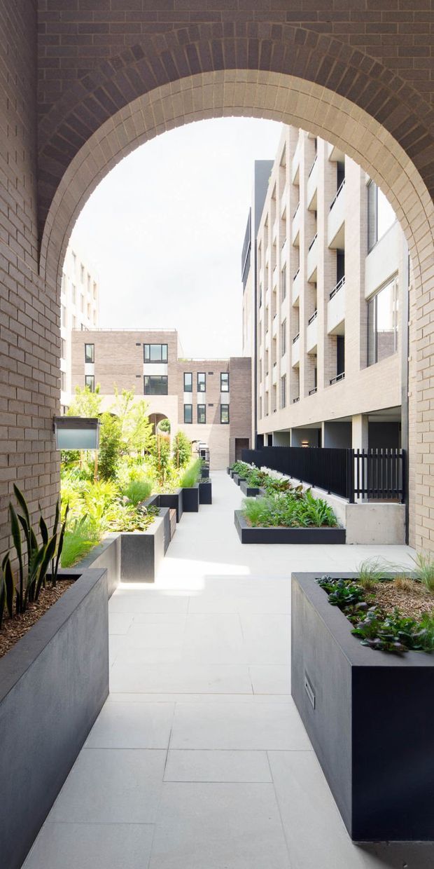 A bright landscaped pedestrian courtyard framed by a large arched brick opening. Pale brick multi-storey residential buildings surround a paved walkway lined with raised dark planters, lush green planting, and black metal fencing. The composition highlights the masonry arch and contemporary apartment courtyard setting.