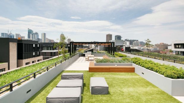 A landscaped rooftop terrace with broad areas of lawn, raised planted beds, timber-edged built-in seating, and outdoor lounge furniture. Dark-framed architectural structures and a dense urban skyline are visible beyond the terrace, creating a contemporary rooftop amenity space.