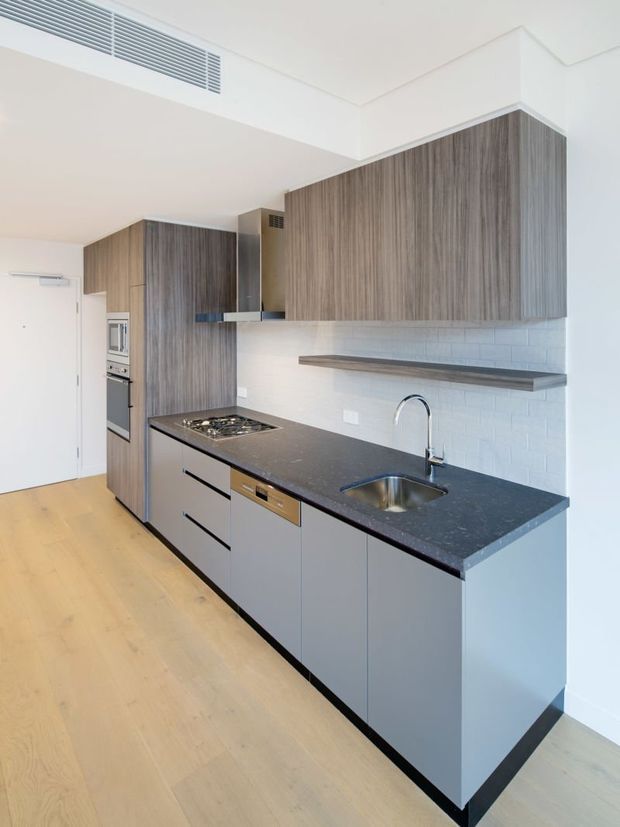 Bright, compact contemporary galley kitchen with a long dark stone-look benchtop, integrated stainless-steel sink and cooktop, grey lower cabinetry, tall vertical-grain timber cabinetry, white painted walls, and a white brick-pattern splashback. Pale timber flooring and concealed appliances create a streamlined, functional fit-out.