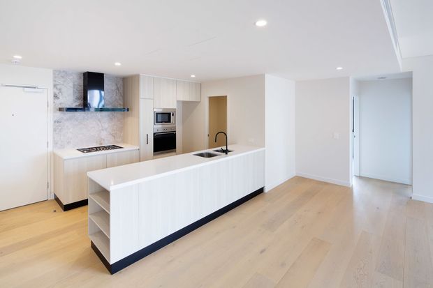 Bright open-plan contemporary kitchen with a long white island, integrated sink and black tap, pale timber cabinetry, built-in oven and microwave, marble-look splashback, and light timber flooring. The kitchen opens into a spacious neutral living or dining area and hallway.