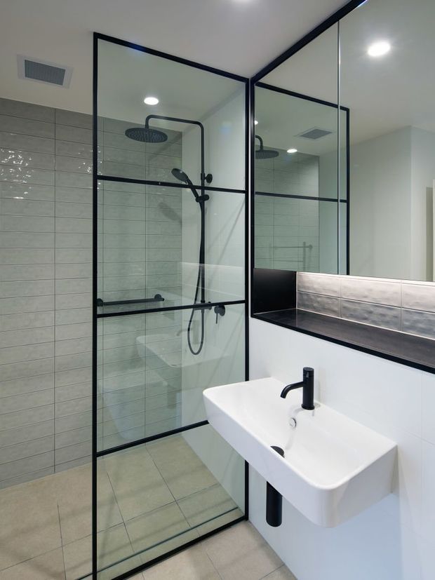 A contemporary bathroom featuring a frameless-style black metal grid shower enclosure with clear glazing, a wall-mounted white basin with matte black tapware, pale grey horizontal wall tiles, light neutral floor tiles, and a large mirrored vanity wall with a dark benchtop. Recessed ceiling lights and a rainfall shower create a clean, modern appearance.