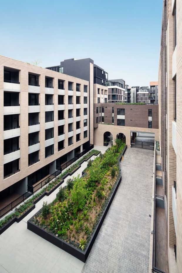 A high-angle view into a landscaped internal courtyard of a contemporary multi-storey apartment development. Pale and dark brick buildings with repetitive recessed windows and balconies frame raised planted garden beds, paved walkways, an arched passageway, and a gated driveway or service access.