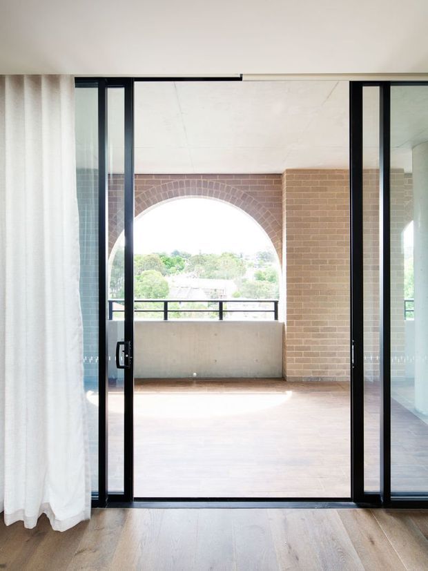 View from an interior room through open black-framed glazed doors onto a covered balcony or terrace. The exterior area features a warm brick wall with a large arched opening, concrete balustrade, dark railing and timber-toned flooring. Light curtains and pale timber flooring frame the foreground, creating a bright, refined indoor-outdoor connection.