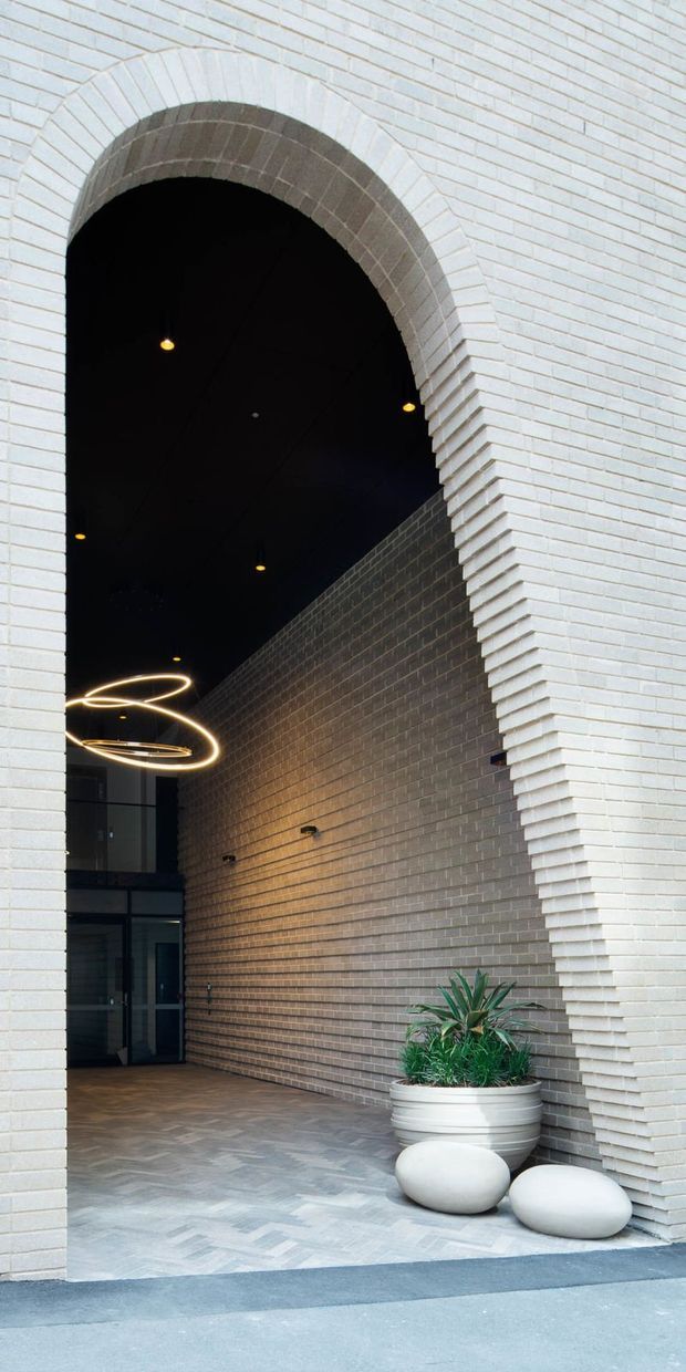 A dramatic arched entryway framed by pale brickwork, leading into a dark, high-ceilinged lobby. The interior features a contrasting darker brick wall, patterned stone or concrete paving, sculptural ring pendant lighting, glass entry doors, and a large planter with greenery and rounded stone forms.