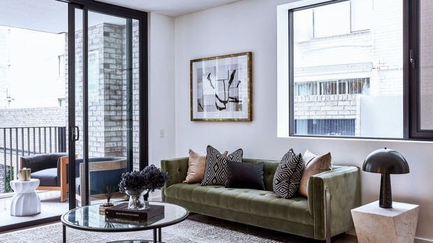 Bright, refined living room with a deep olive-green upholstered sofa, patterned and neutral cushions, a round glass coffee table, sculptural black table lamp, framed abstract artwork, and large black-framed windows and glazing opening toward a balcony. The white walls and abundant daylight create a polished contemporary apartment interior.