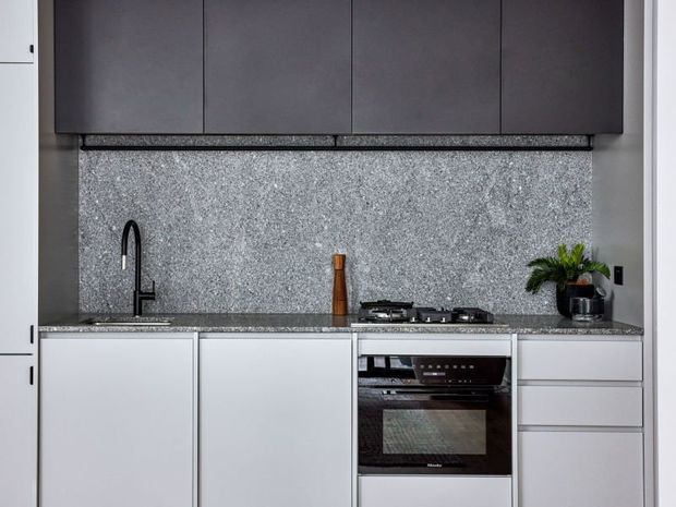Compact modern kitchen featuring matte dark grey overhead cabinetry, pale grey lower cabinetry, a speckled grey stone benchtop and full-height splashback, integrated oven, gas cooktop, and a black curved gooseneck tap. A small potted plant adds greenery.