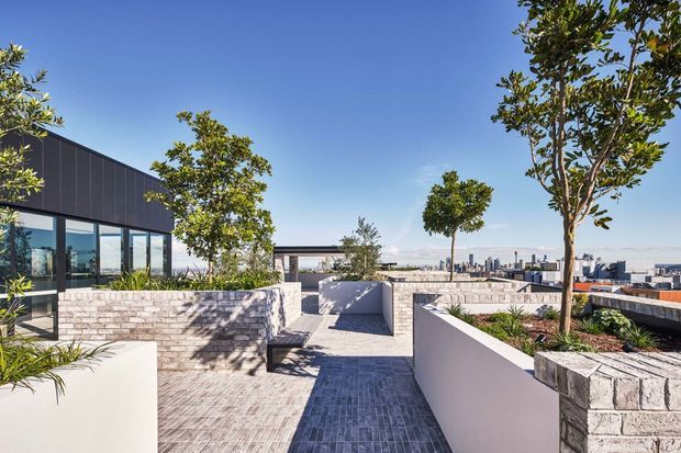 A contemporary rooftop terrace with paved walkways, raised masonry planters, integrated landscaping, and a dark-clad glazed pavilion. The elevated outdoor space overlooks a dense city skyline under clear blue skies.